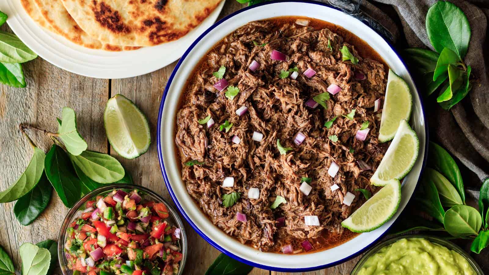 A bowl filled with shredded beef, chopped onions, and cilantro, garnished with lime wedges. Surrounding the bowl are tortillas, guacamole, and salsa on a wooden table.