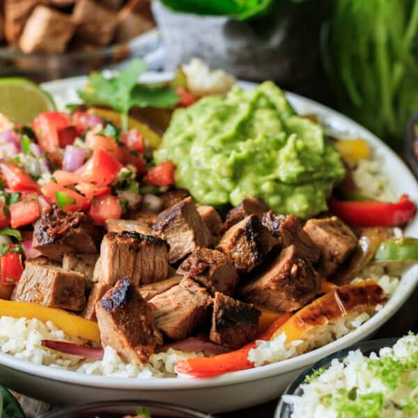 A bowl containing grilled chicken, beef, sliced bell peppers, pico de gallo, guacamole, and rice. In the background, small bowls hold guacamole and assorted vegetables.