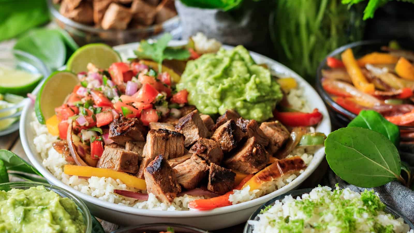 A bowl containing grilled diced steak, guacamole, pico de gallo, sliced bell peppers, and rice is placed at the center. Surrounding the bowl are side dishes including sliced avocado, tortilla chips with salsa in a small bowl, and lime wedges.