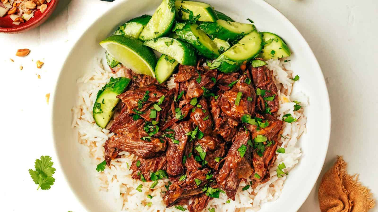 A bowl containing white rice, shredded beef, chopped cilantro, sliced cucumbers, and lime wedges.