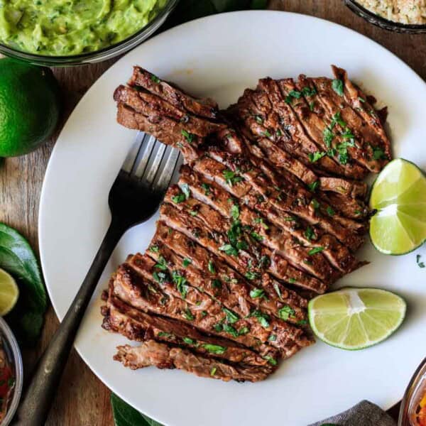 A plate with grilled, sliced steak garnished with chopped herbs and lime wedges, surrounded by bowls of guacamole, salsa, quinoa, and sautéed bell peppers.