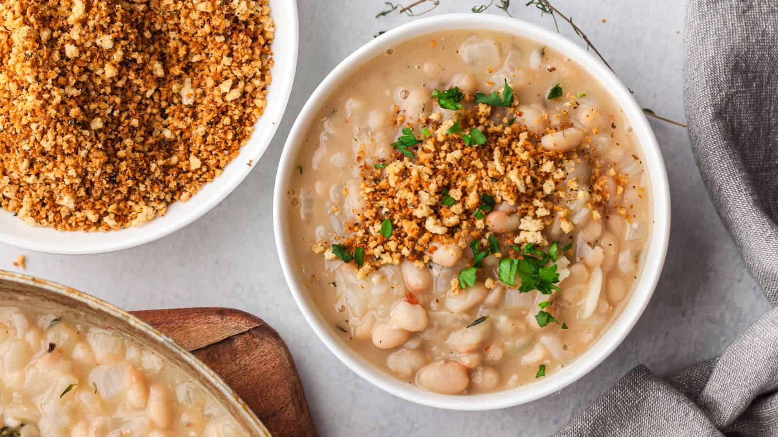 A bowl of creamy white bean stew topped with chopped parsley and crispy breadcrumbs, beside a smaller bowl filled with extra breadcrumbs.