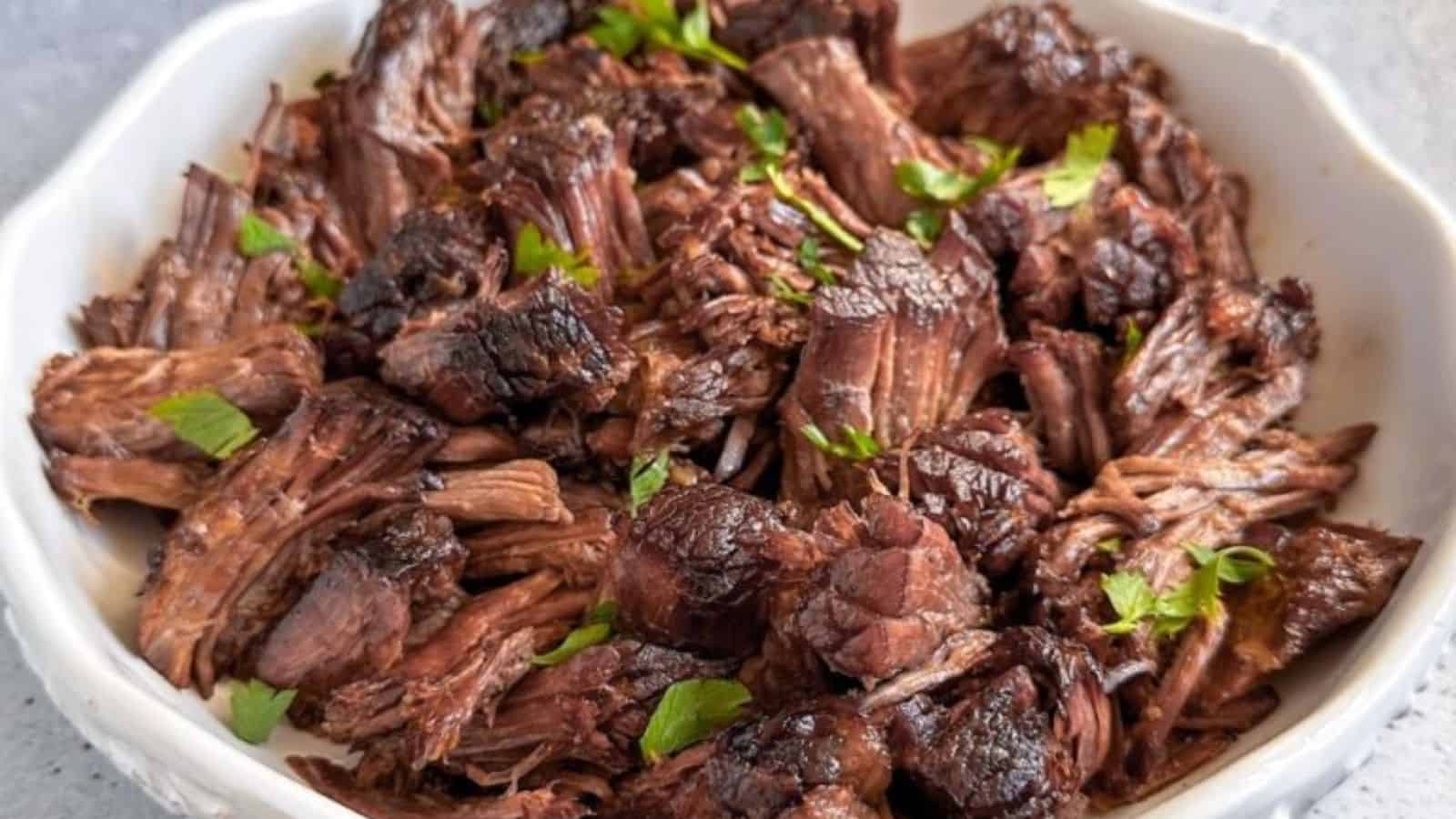 A white bowl containing shredded, cooked beef topped with chopped parsley.