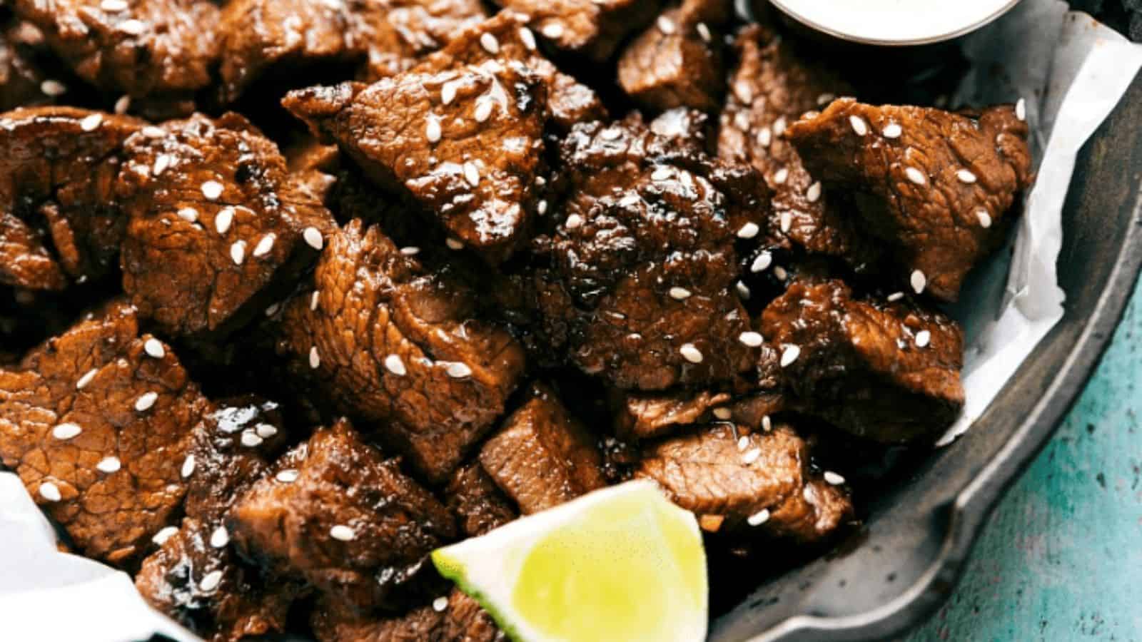 Cooked beef steak bites sprinkled with sesame seeds, accompanied by a lime wedge and a small container of dipping sauce, all served in a bowl.