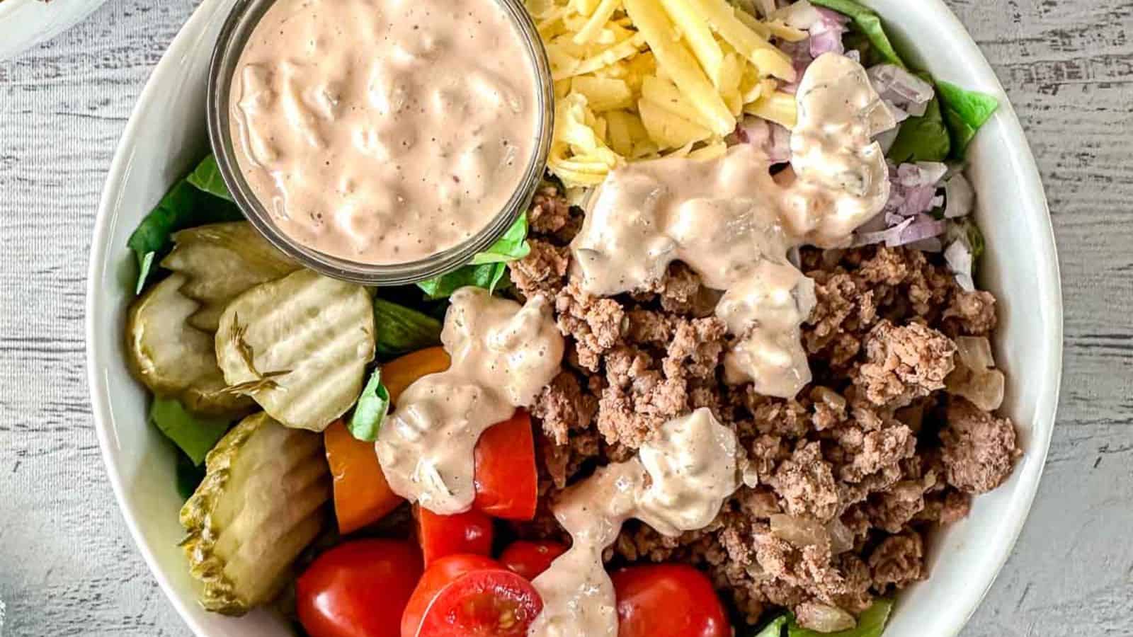 A bowl containing ground beef, chopped tomatoes, pickles, shredded cheese, onions, lettuce, and a small container of creamy dressing.