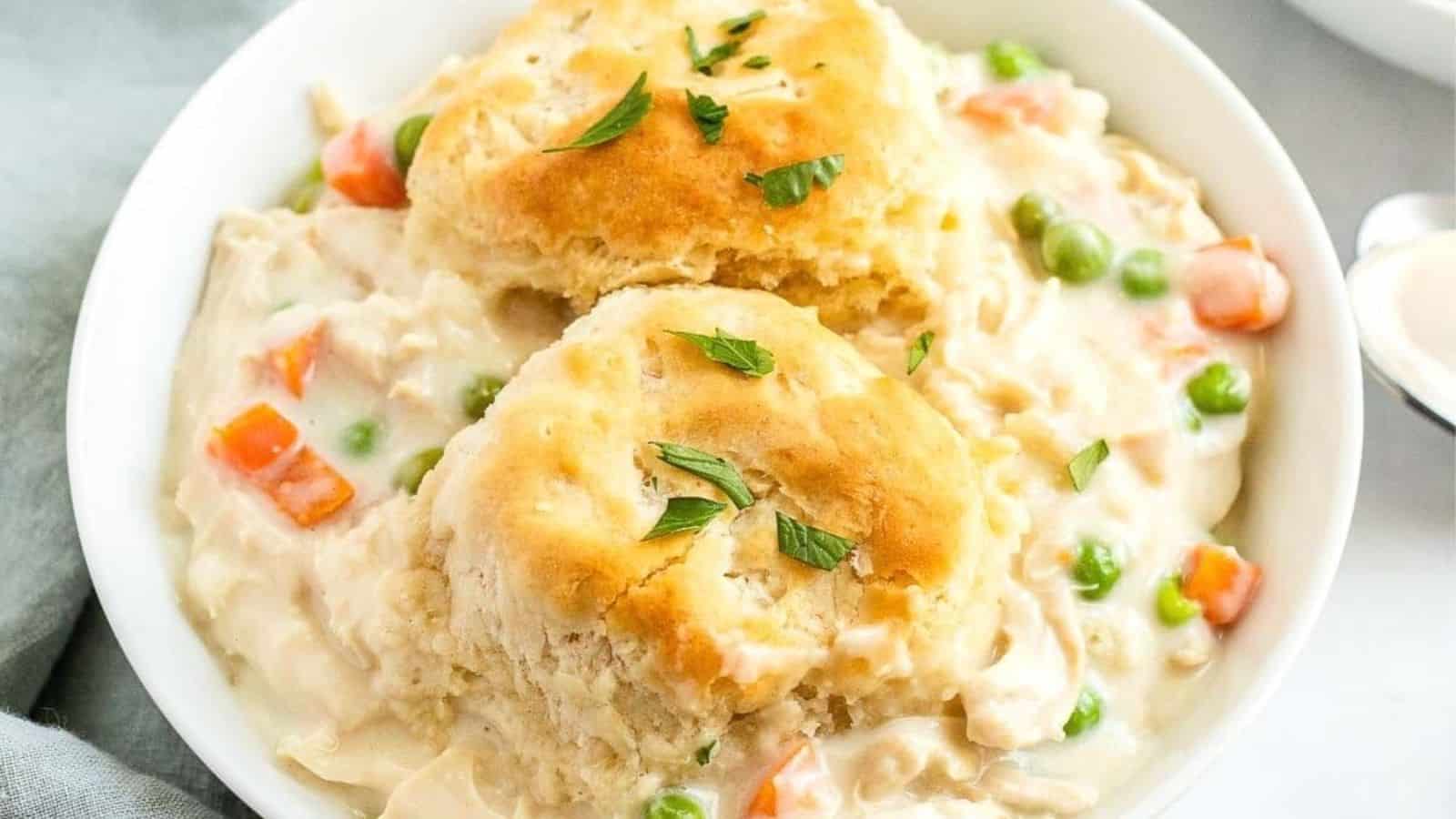 A white bowl containing creamy chicken and vegetable stew, with two golden-brown biscuits on top and a sprinkle of chopped parsley.