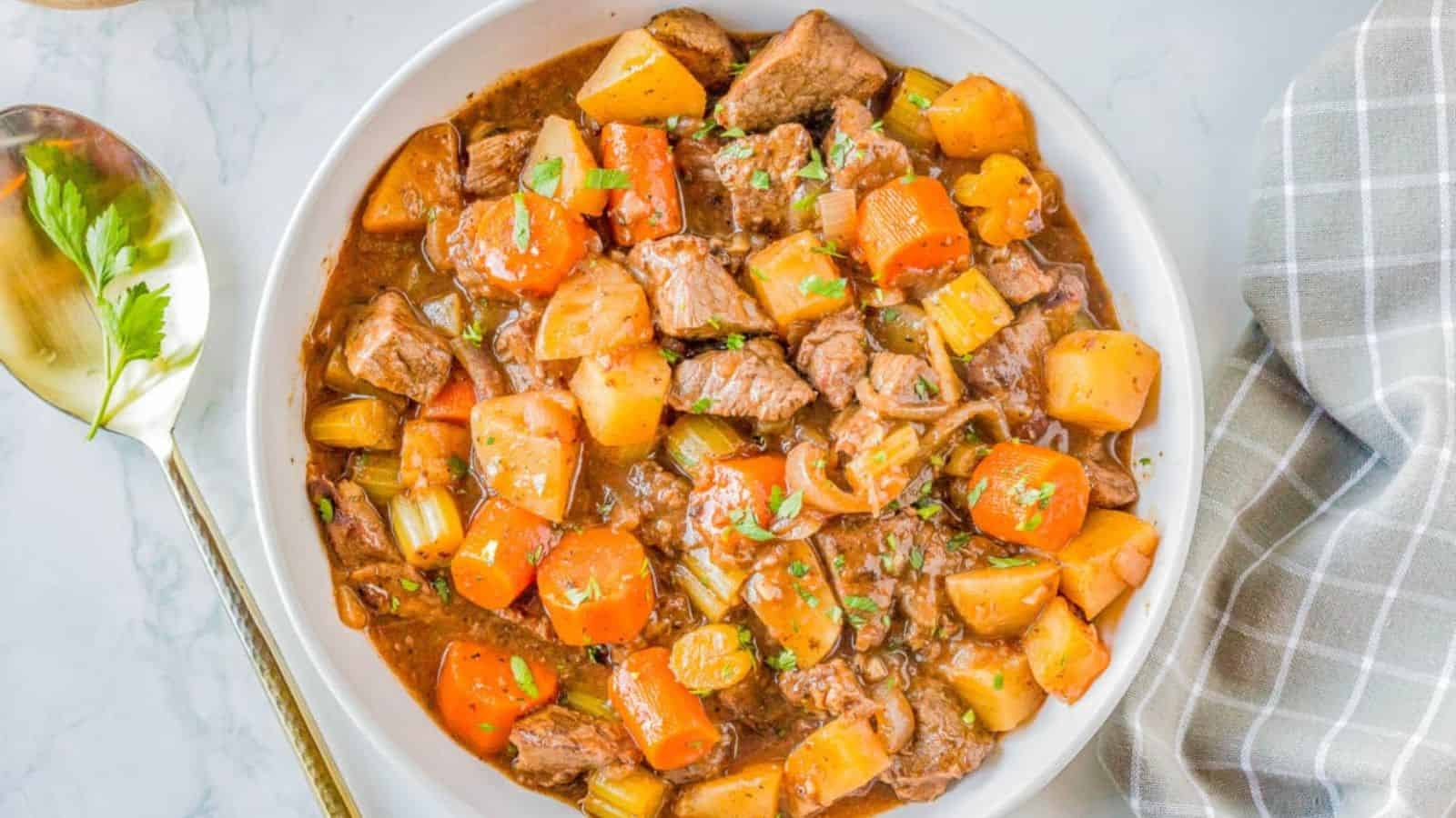 A bowl of beef stew containing chunks of beef, carrots, potatoes, and celery in a thick brown broth, topped with chopped herbs. A spoon and a gray plaid napkin are placed beside the bowl.