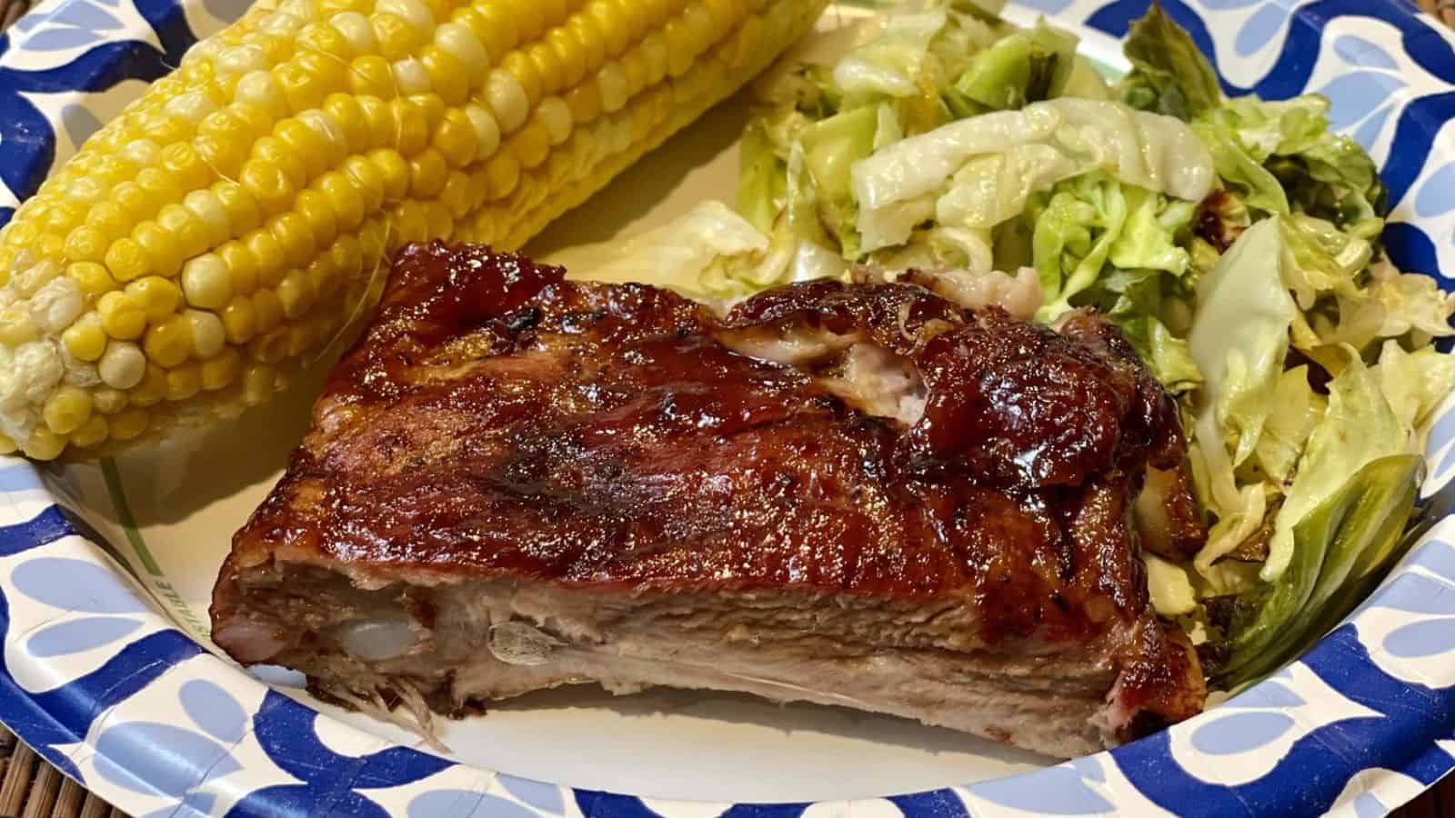 Barbecued ribs, corn on the cob, and cooked cabbage arranged on a blue and white paper plate.