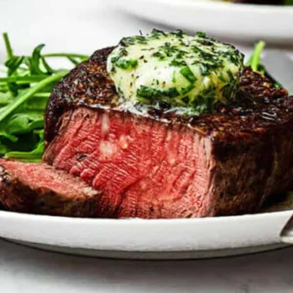 A medium-rare steak with herb butter, fresh arugula, a white plate, a fork, and a knife.