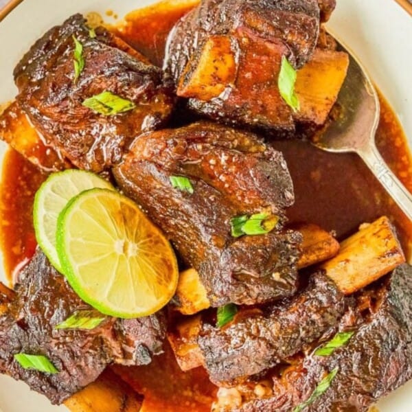 Braised beef short ribs in sauce on a plate, topped with sliced green onions and lime wedges, with a silver spoon beside the plate.
