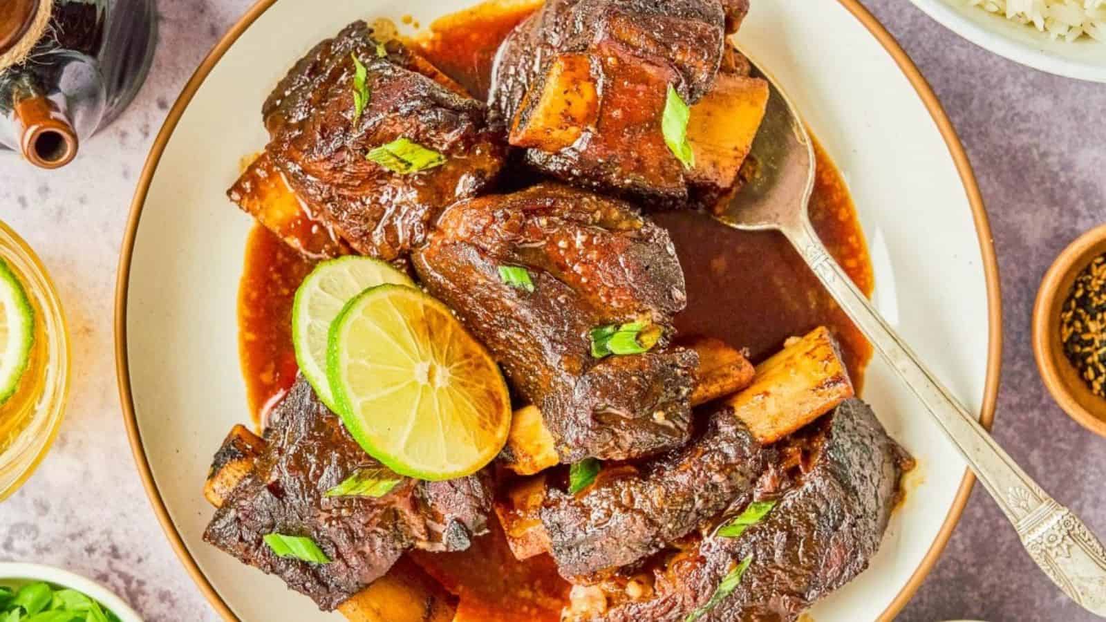 Braised beef short ribs in sauce on a plate, topped with sliced green onions and lime wedges, with a silver spoon beside the plate.