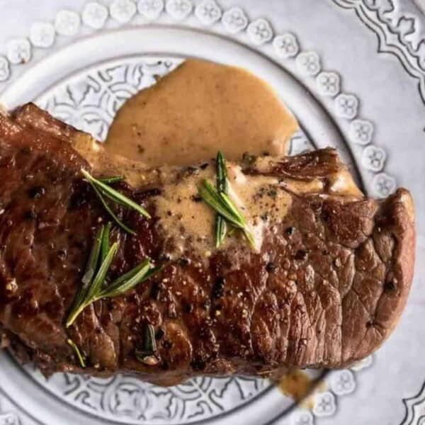 The image features a grilled steak with a sear, topped with rosemary and sauce, presented on a decorative white plate.