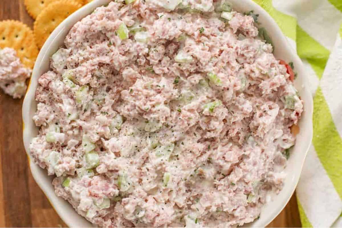 A bowl of creamy ham salad mixed with chopped celery and mayonnaise, garnished with black pepper, sits on a wooden surface with round crackers nearby.
