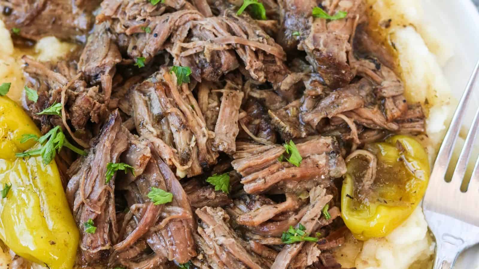 Shredded beef pot roast on a bed of mashed potatoes, topped with pepperoncini peppers and chopped parsley.