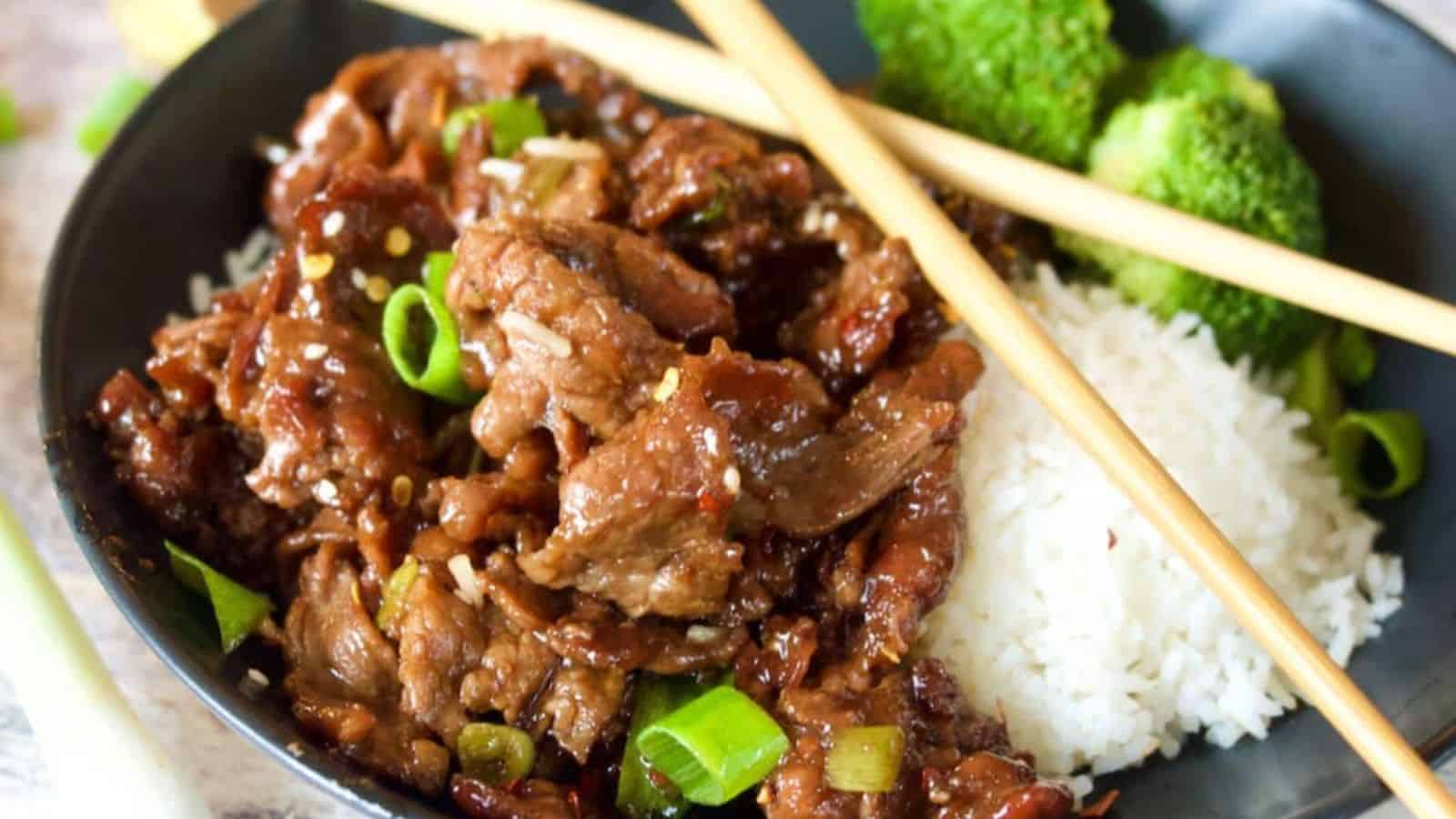 A black bowl containing stir-fried beef, white rice, broccoli florets, and sliced green onions; chopsticks rest on the bowl.
