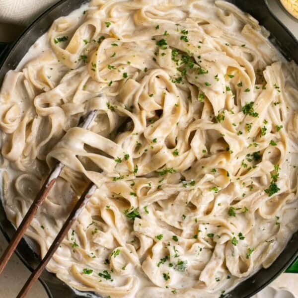 A skillet containing fettuccine Alfredo garnished with chopped parsley, alongside serving utensils and a bowl of grated cheese.
