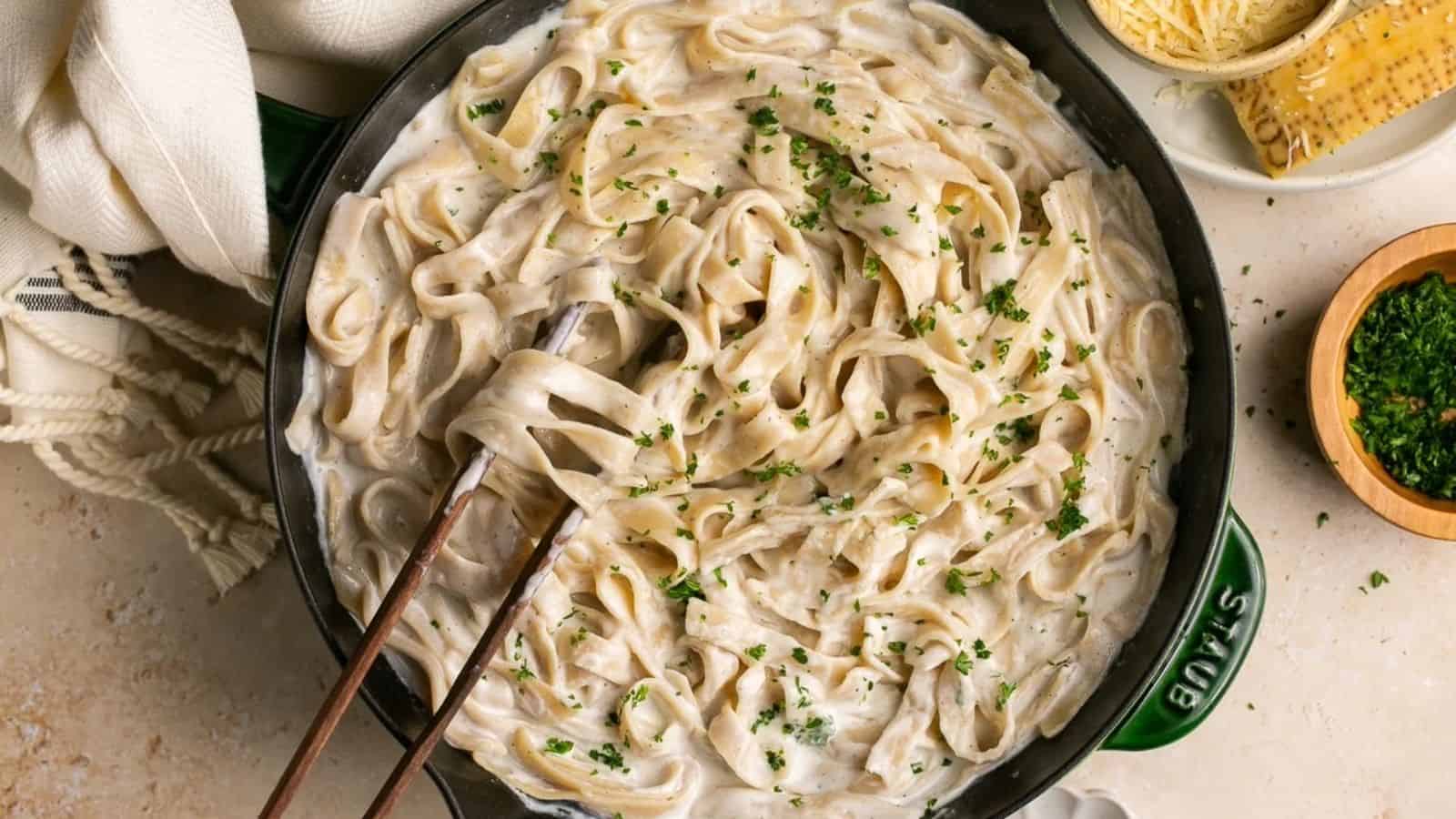 A skillet containing fettuccine Alfredo garnished with chopped parsley, alongside serving utensils and a bowl of grated cheese.