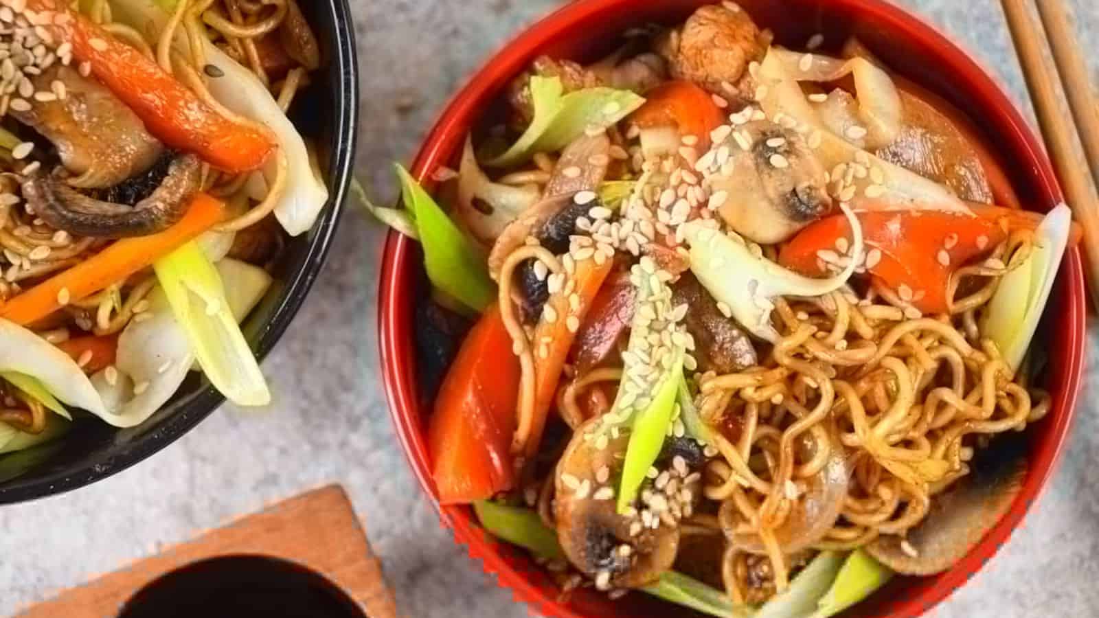 Two bowls filled with stir-fried noodles, mushrooms, red bell peppers, green onions, and sesame seeds. Chopsticks rest beside the bowls, and a small dish of soy sauce is placed nearby.