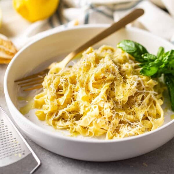A plate of fettuccine pasta topped with grated cheese and herbs, garnished with fresh basil, with a fork resting on the side.
