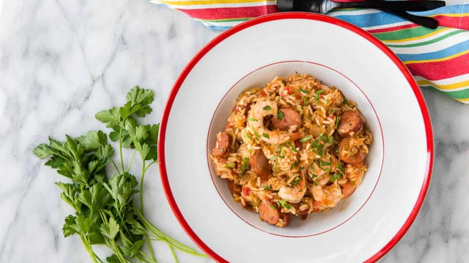 A bowl filled with jambalaya containing shrimp and sausage, garnished with parsley, is placed on a marble surface. Nearby are sprigs of fresh parsley and a striped cloth napkin.