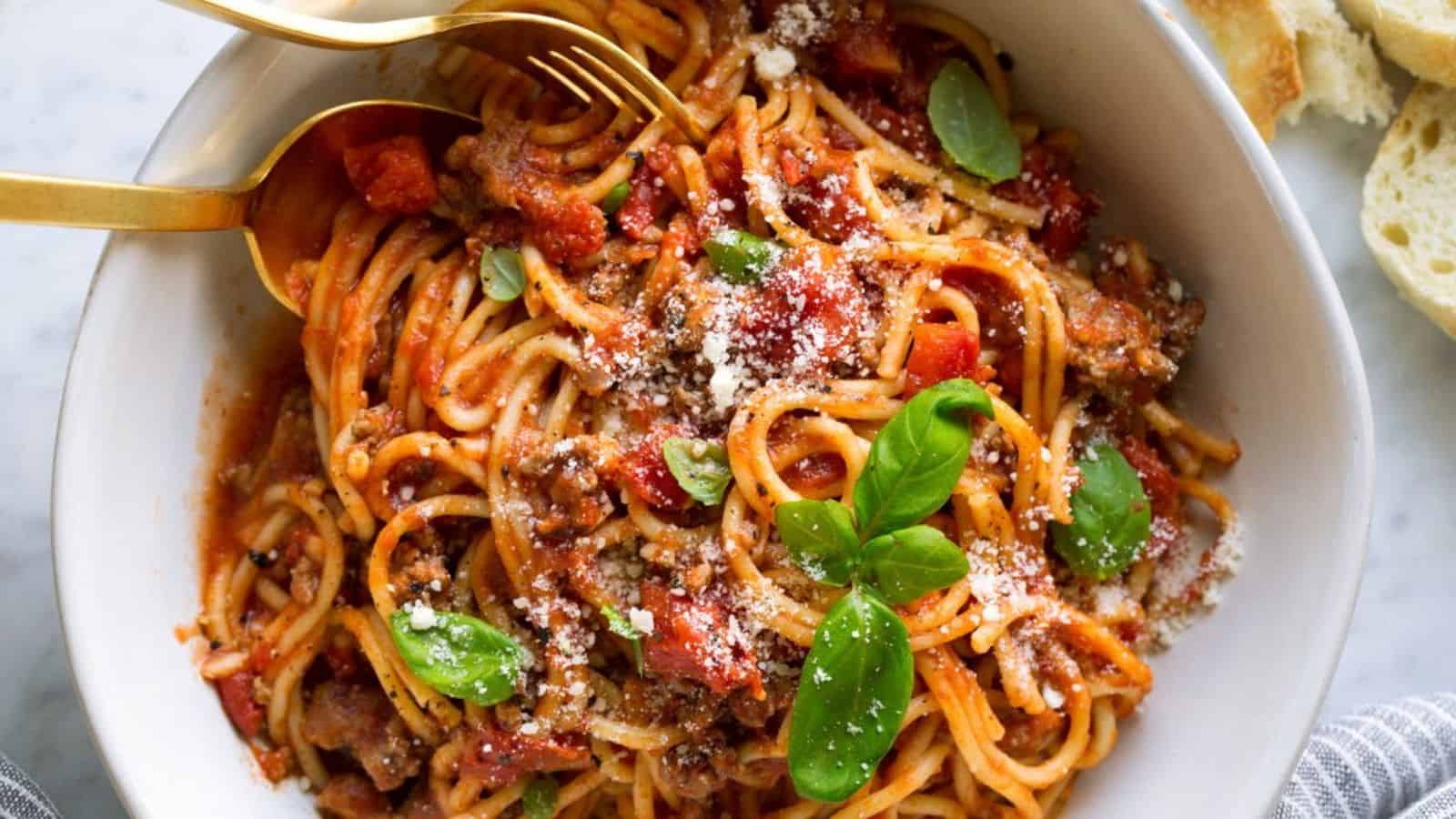 A bowl of spaghetti topped with tomato meat sauce, grated cheese, and fresh basil leaves; gold utensils and slices of bread are placed nearby.