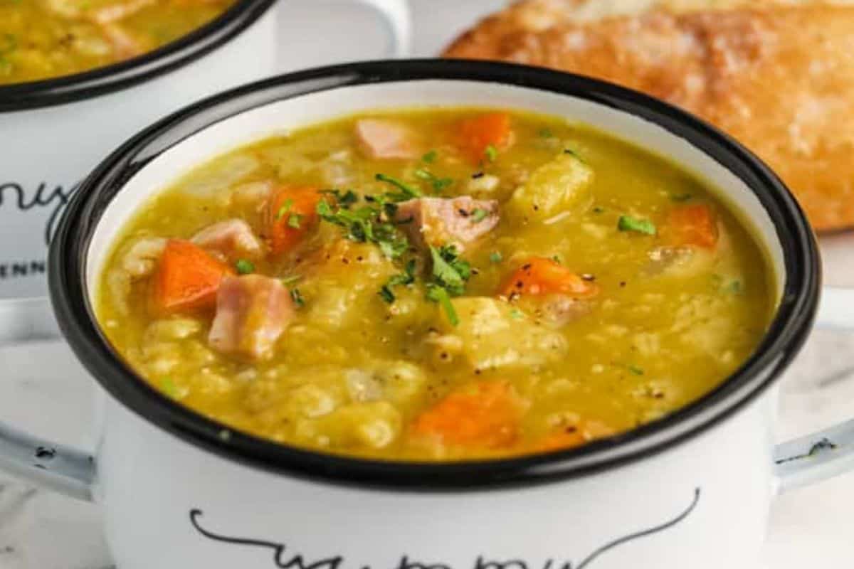 A white bowl filled with chunky split pea soup, featuring pieces of ham, carrots, and potatoes, garnished with chopped herbs. A piece of bread is visible in the background.