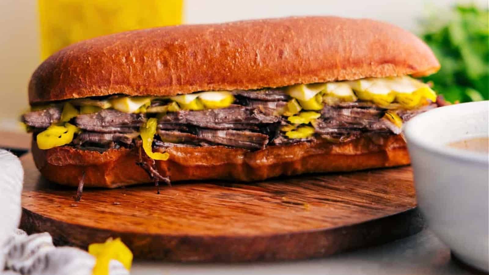 A hoagie roll filled with roast beef, sliced peppers, and pickles sits on a wooden board beside a white bowl.