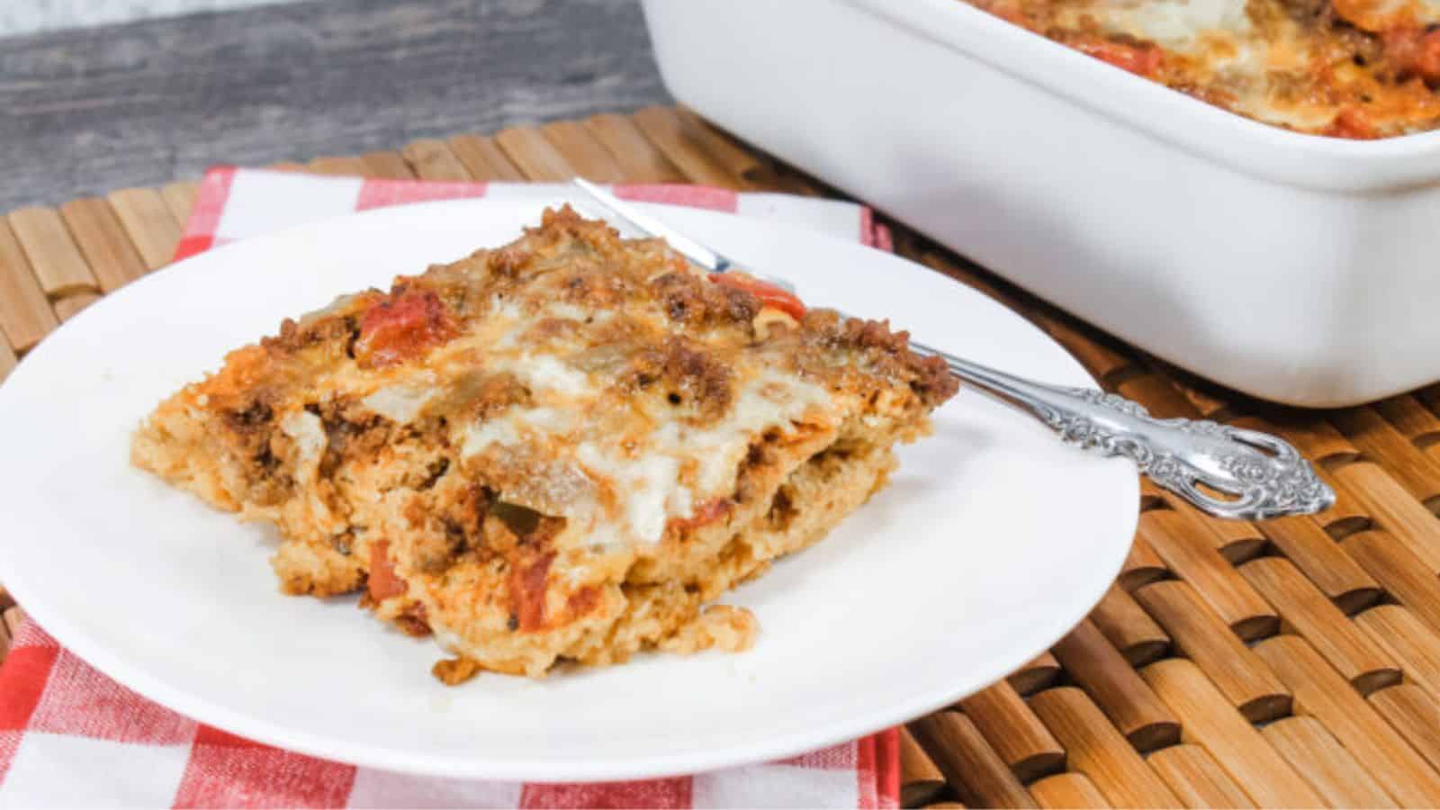 A slice of casserole topped with melted cheese and tomato sauce on a white plate with a fork, next to a glass baking dish containing more casserole.