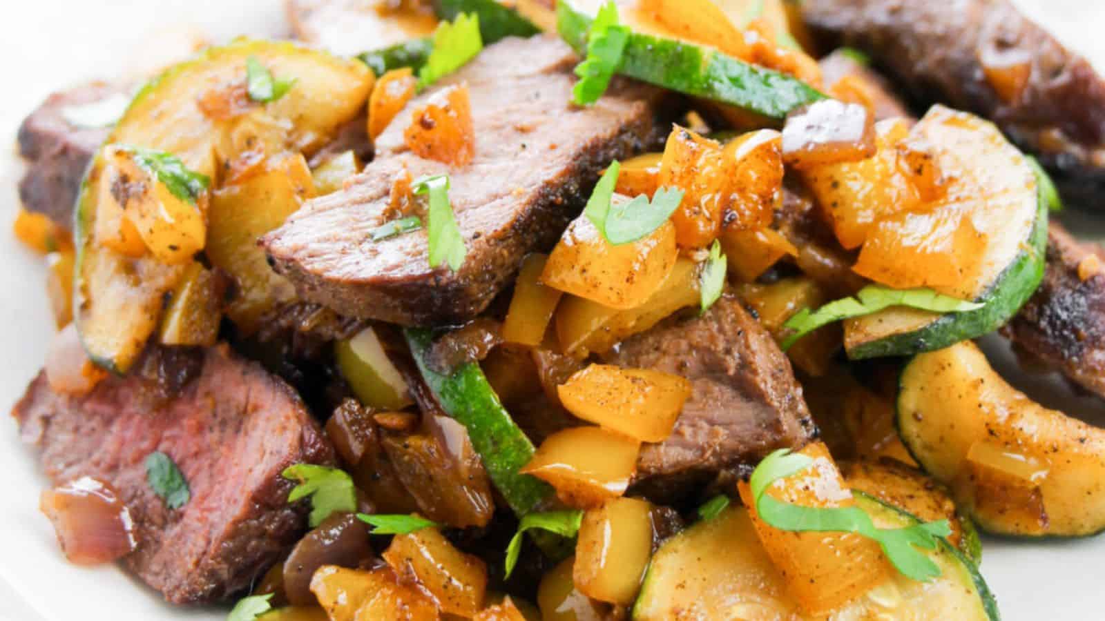 Sliced steak, zucchini, yellow bell peppers, and onions garnished with fresh herbs on a white plate.