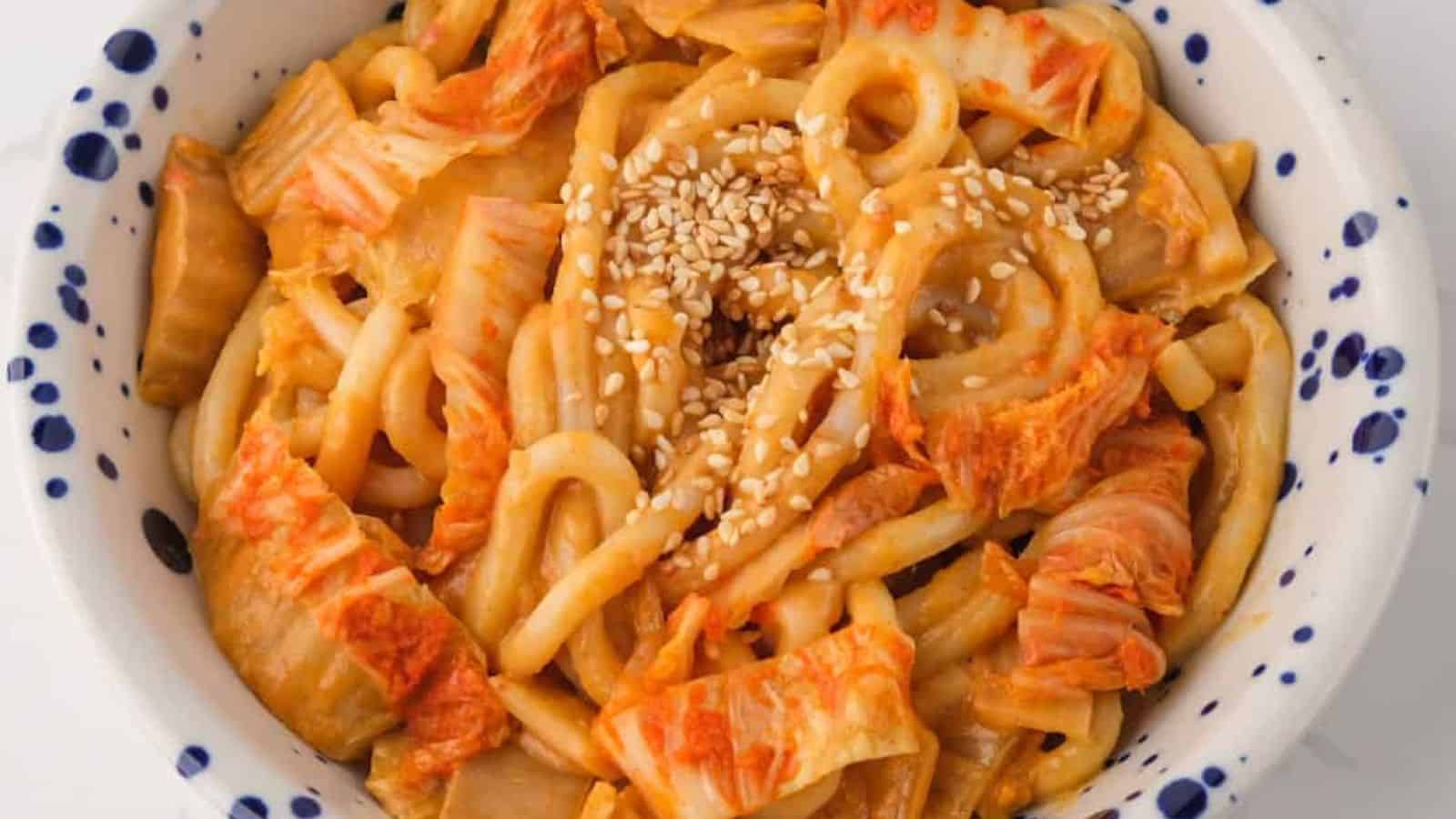 A white bowl with blue spots filled with udon noodles, kimchi, and a sprinkling of sesame seeds.