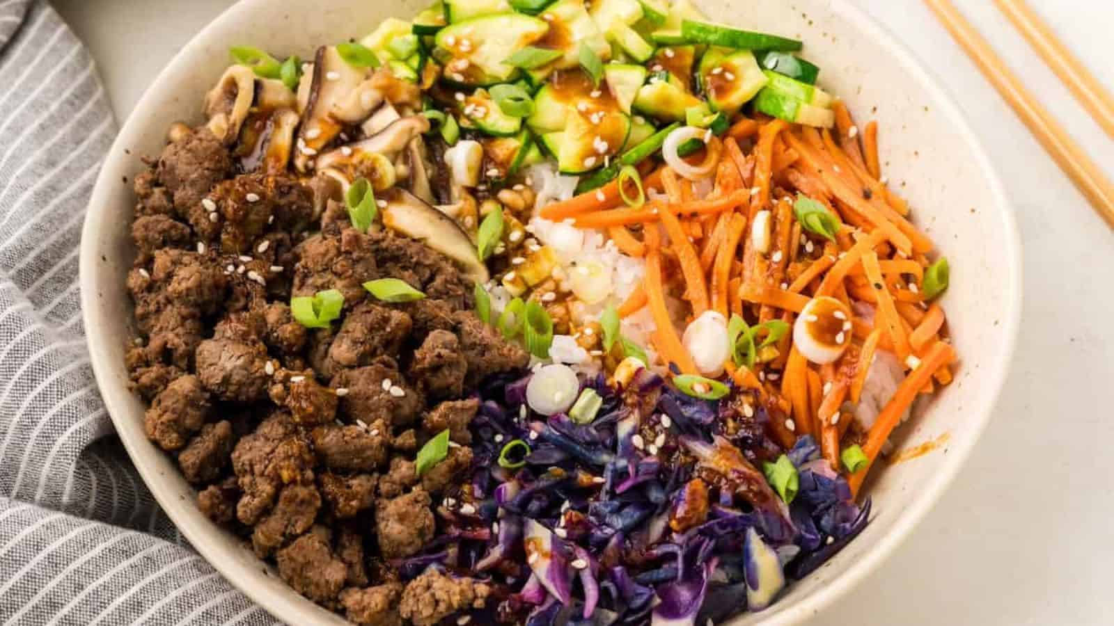 A bowl containing ground beef, shredded carrots, sliced cucumber, shiitake mushrooms, chopped green onions, red cabbage, rice, sesame seeds, and sauce.