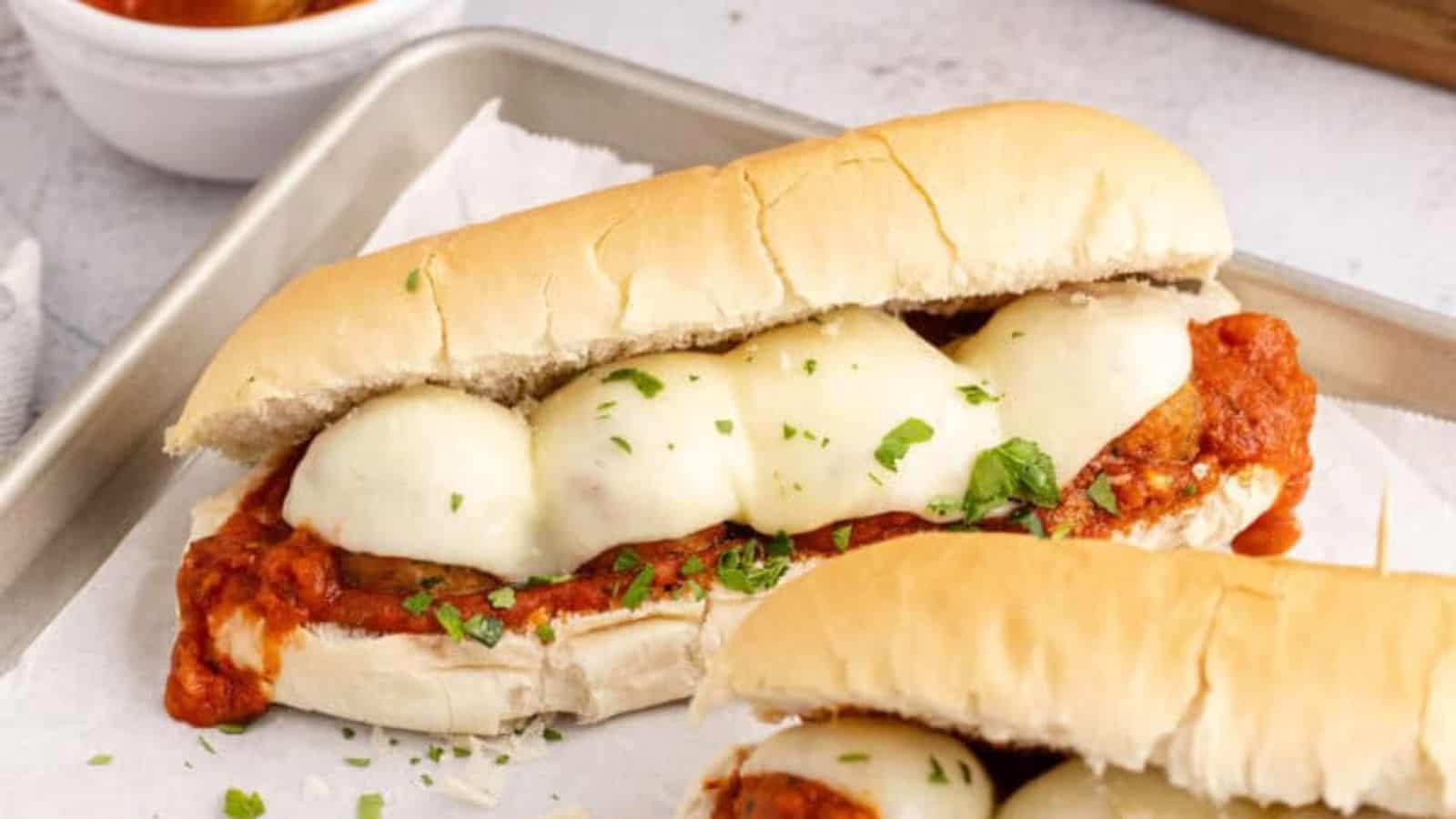 A sandwich filled with meatballs, melted mozzarella cheese, and tomato sauce, garnished with chopped herbs, served on a metal tray lined with parchment paper.