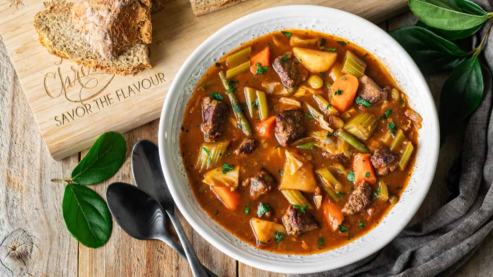 A bowl filled with beef stew containing chunks of potatoes, carrots, and celery, accompanied by slices of bread on a wooden board and two spoons placed nearby.