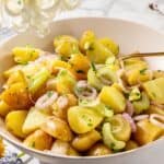 A bowl of potato salad with sliced onions and celery, garnished with herbs. A silver spoon is in the mix, surrounded by delicate flowers on a white surface.