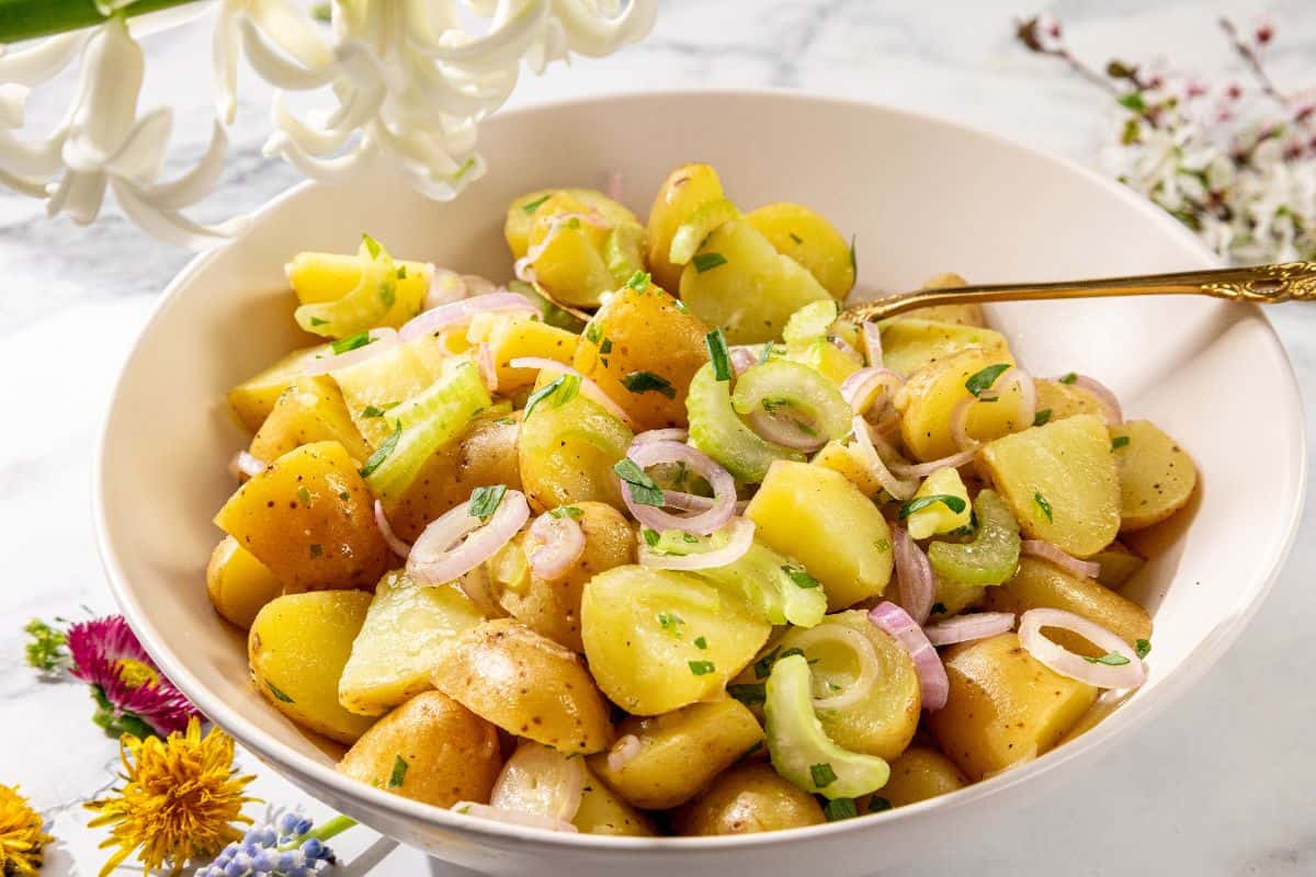A bowl of potato salad with sliced onions and celery, garnished with herbs. A silver spoon is in the mix, surrounded by delicate flowers on a white surface.