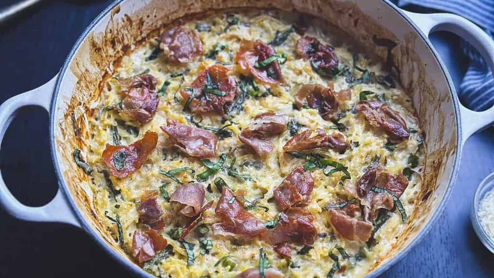 A casserole dish containing a creamy mixture, topped with browned prosciutto slices and sprinkled with chopped green herbs.
