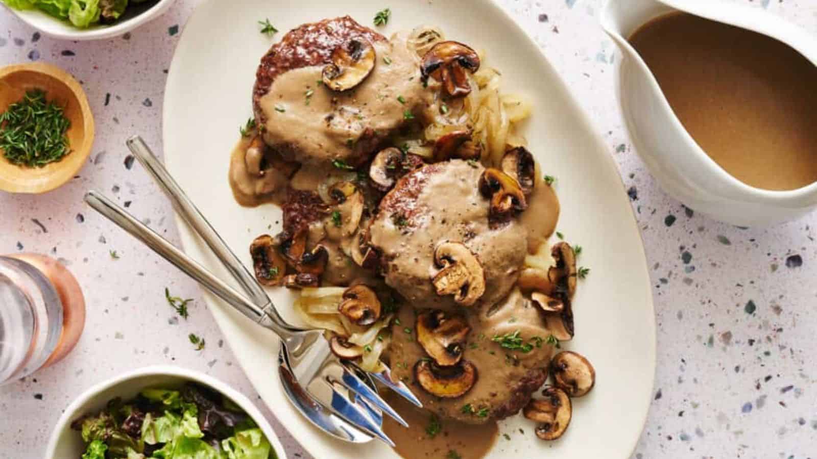 An oval plate holding two Salisbury steaks covered with brown mushroom gravy, sautéed mushrooms, onions, and herbs. Beside the plate are a small jug filled with extra gravy and a bowl of salad.
