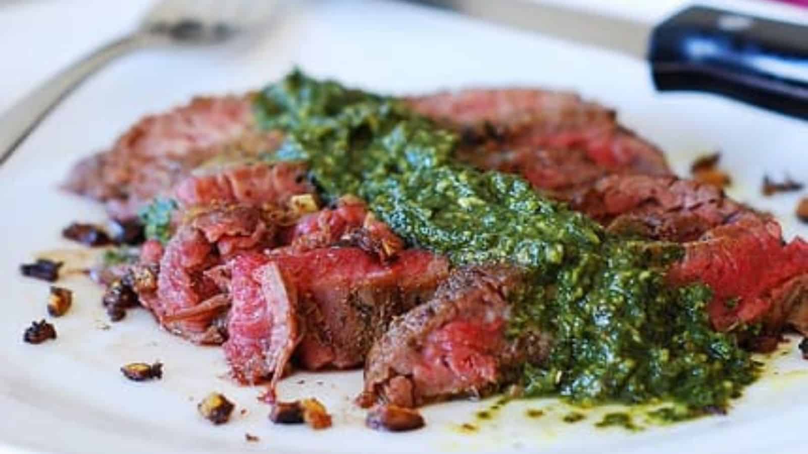 Slices of medium-rare steak topped with green chimichurri sauce, garnished with pieces of roasted garlic on a white plate.