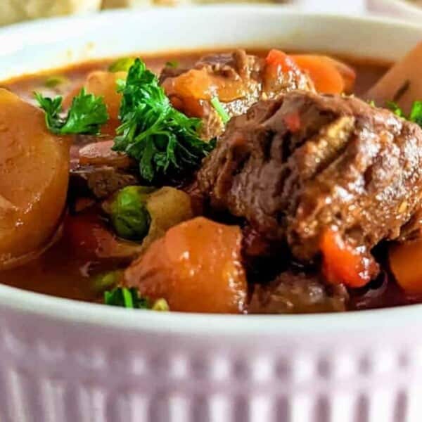 Chunks of beef, potatoes, carrots, and peas in a pink ribbed bowl, garnished with parsley.