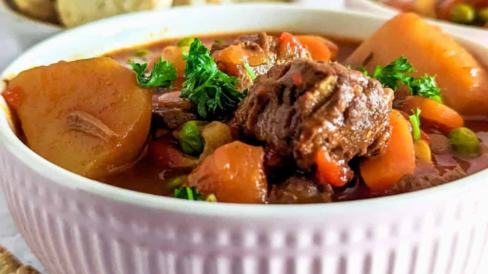 Chunks of beef, potatoes, carrots, and peas in a pink ribbed bowl, garnished with parsley.