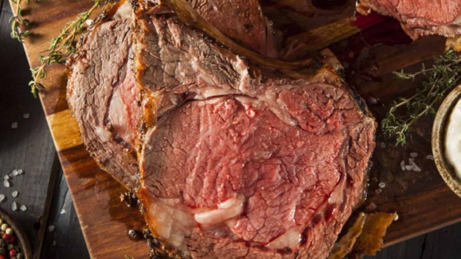 Slices of medium-rare prime rib roast with visible marbling are arranged on a wooden cutting board, surrounded by sprigs of fresh herbs.