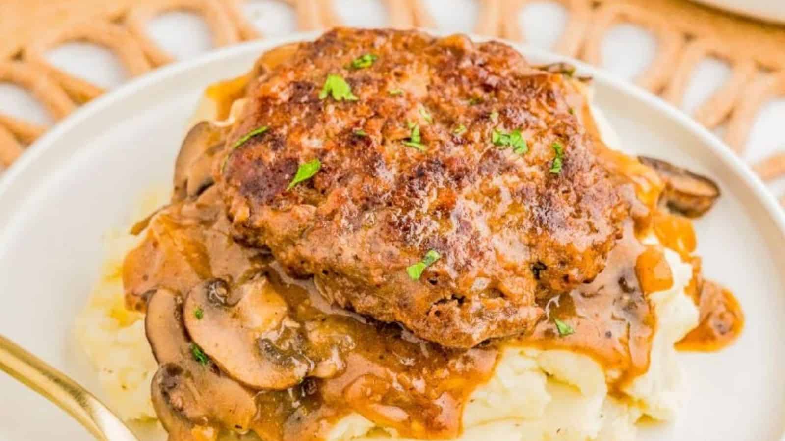 A hamburger steak topped with brown mushroom gravy rests on mashed potatoes, garnished with chopped parsley.