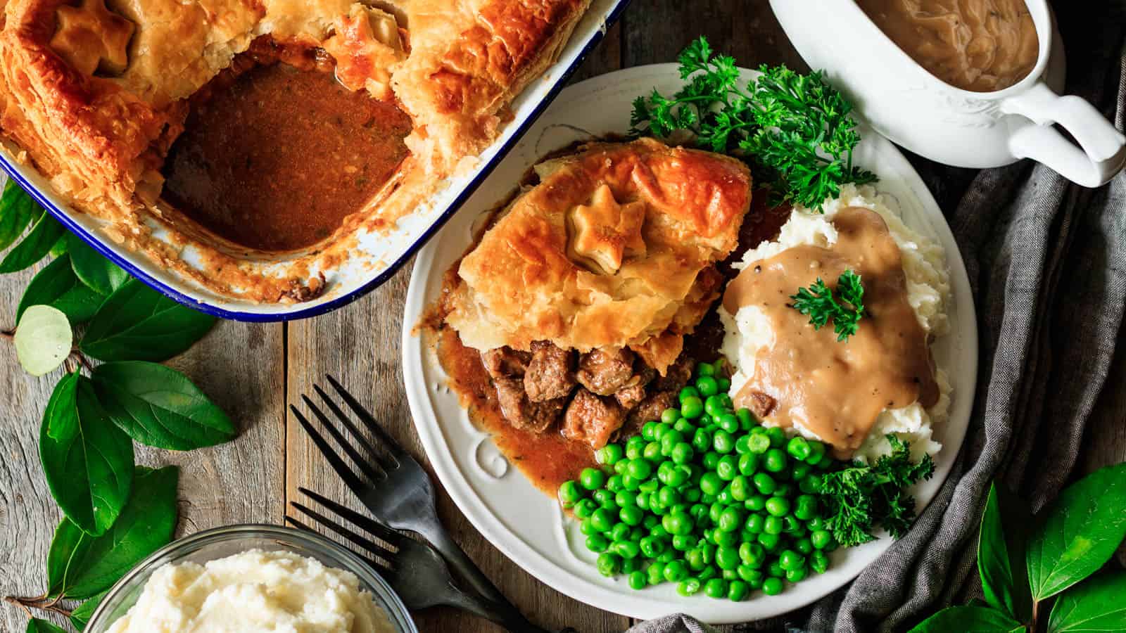 A plate containing beef pot pie with a golden crust, mashed potatoes topped with brown gravy, green peas, and parsley garnish. Nearby is a baking dish of pot pie with one slice removed.