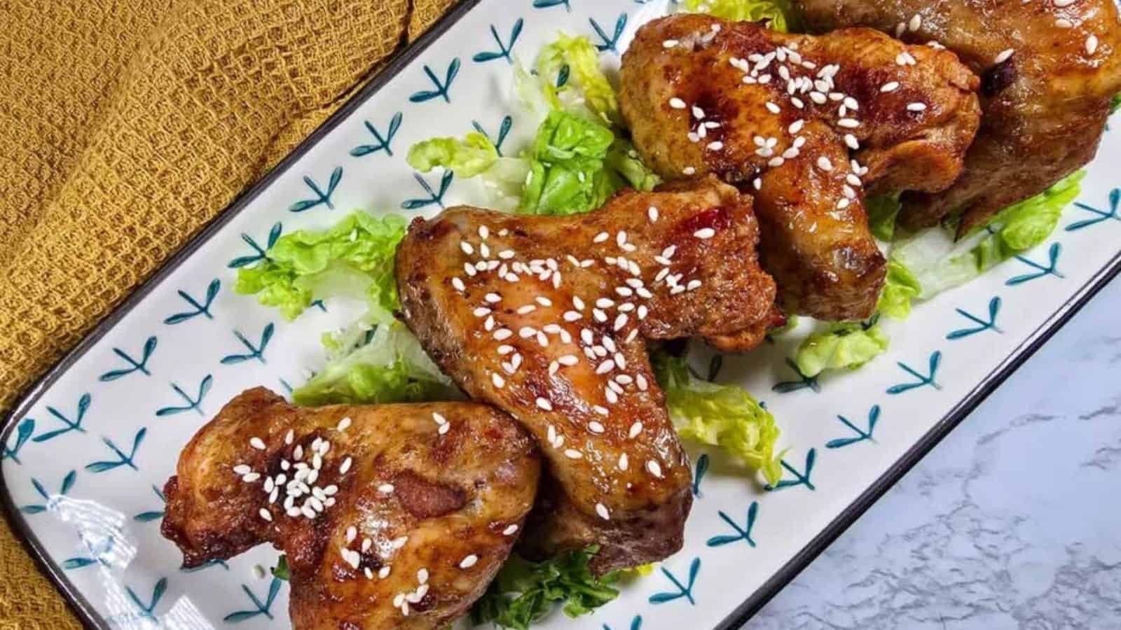 Three glazed chicken wings topped with sesame seeds are placed on shredded lettuce on a rectangular plate.