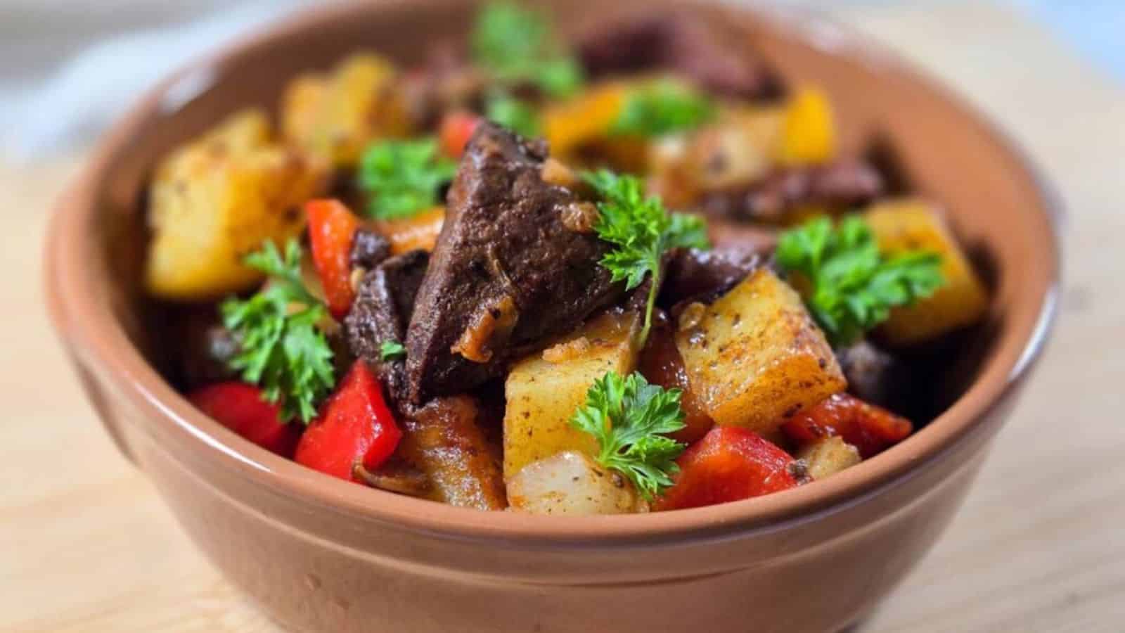 A brown bowl containing a stew with chunks of beef, potatoes, red bell peppers, and herbs, topped with fresh parsley.