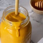 A glass jar containing a thick yellow liquid with a wooden stick inside. In the background, a bowl with a honey dipper resting across it.