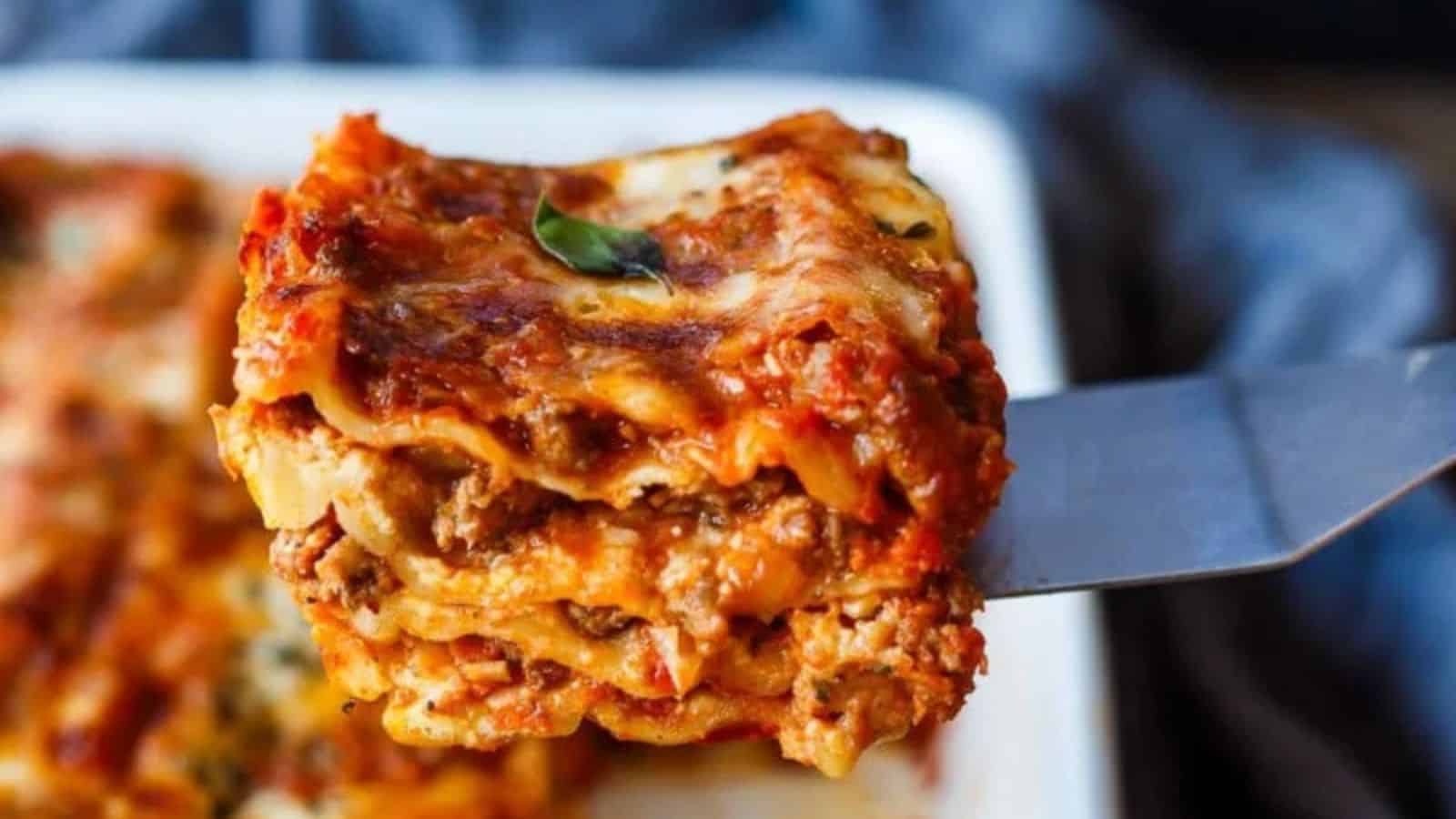 A slice of lasagna with visible layers of pasta, meat, cheese, and tomato sauce is held above a baking dish on a spatula.