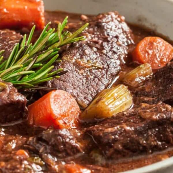 Chunks of beef, carrots, and celery in a thick brown broth, garnished with a sprig of rosemary inside a bowl.