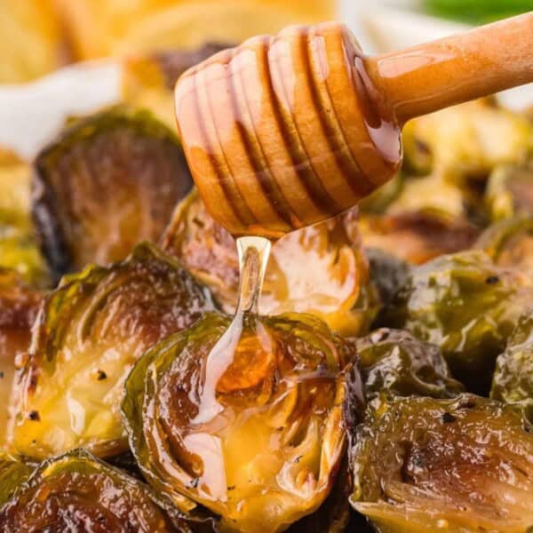 A wooden honey dipper drizzles golden honey over roasted Brussels sprouts, surrounded by warm bread slices in the background.
