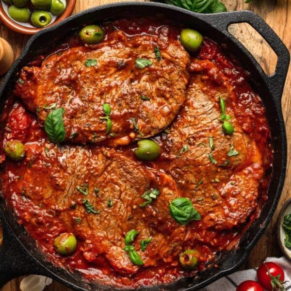 A cast iron skillet with beef in tomato sauce, topped with green olives and basil, next to sliced bread and small bowls of herbs.